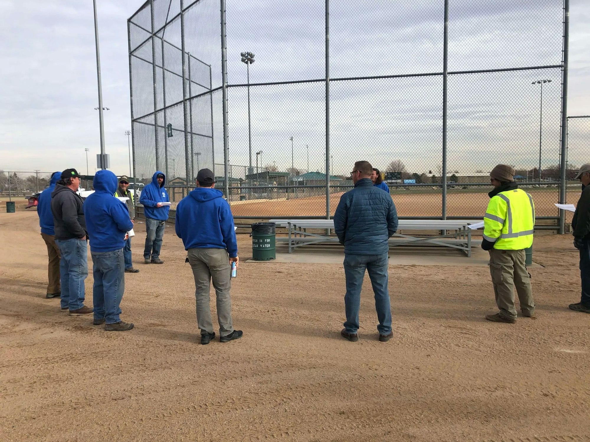 Group of individuals standing near a baseball diamond fence and bleachers.