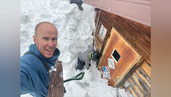 A man takes a selfie with another person shoveling snow by a cabin.