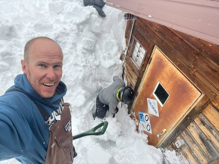 A man takes a selfie with another person shoveling snow by a cabin.