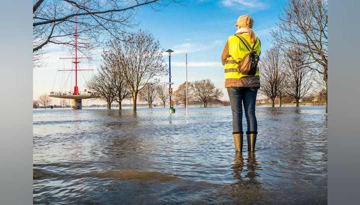 Person in high-vis jacket stands in a flooded area with bare trees, submerged signs, and a ship structure in the background.