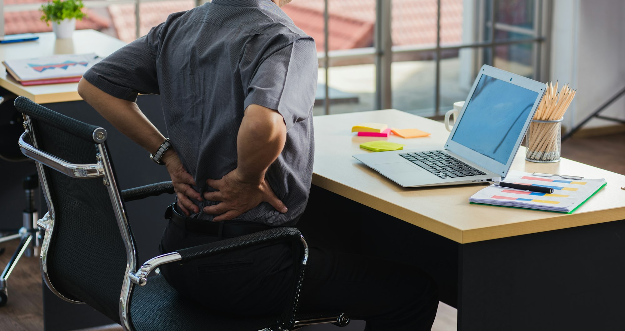 A person in an office is sitting at a desk with a laptop, looking uncomfortable while holding their lower back.
