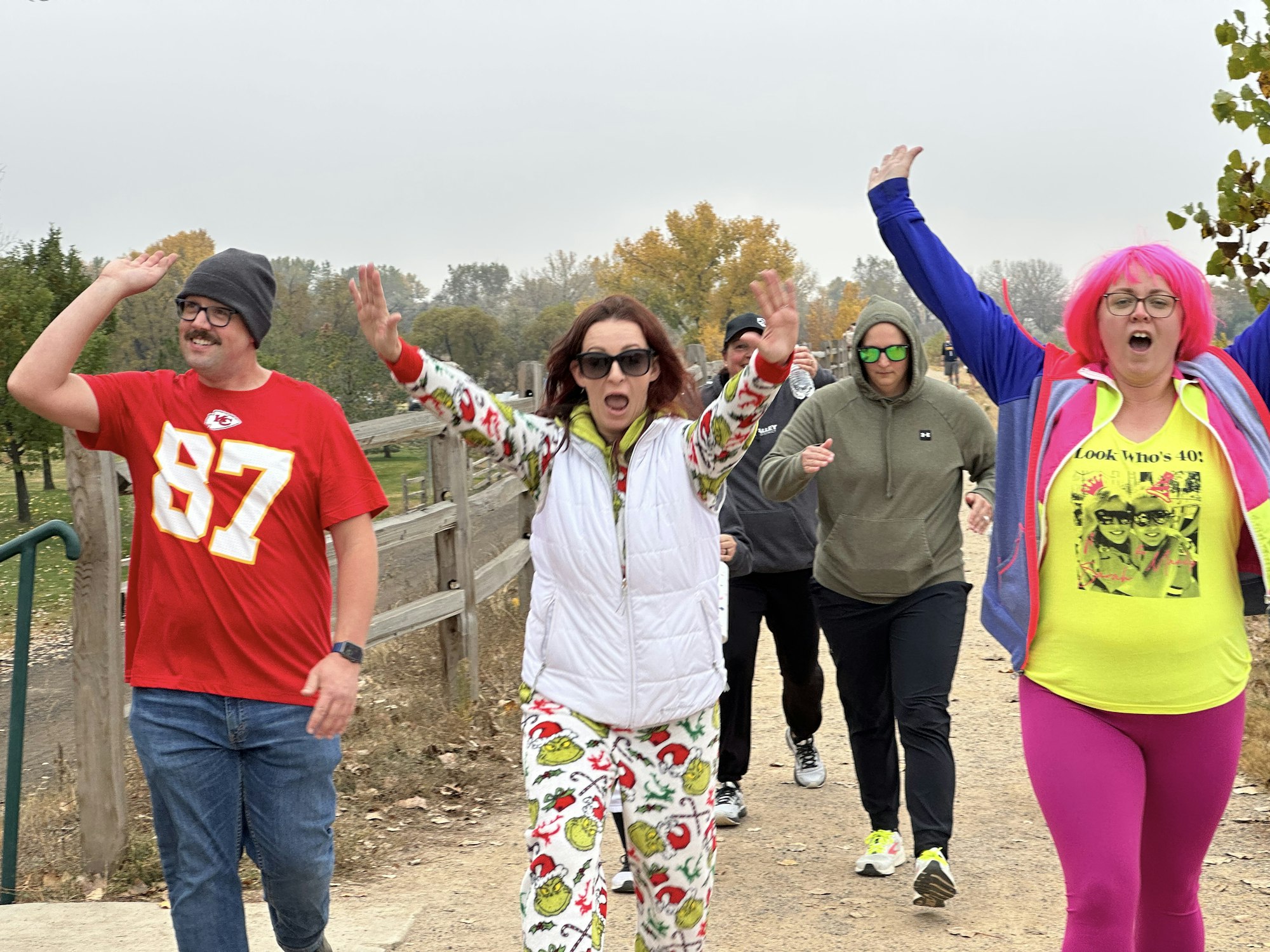 A group of people jogging, arms raised, wearing colorful clothing and glasses, outside on a pathway with trees in the background.