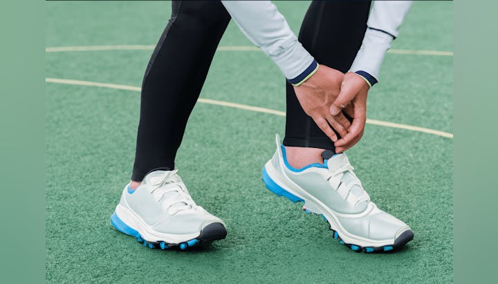 Person holding their ankle while standing on a green surface, wearing sports shoes and black leggings.