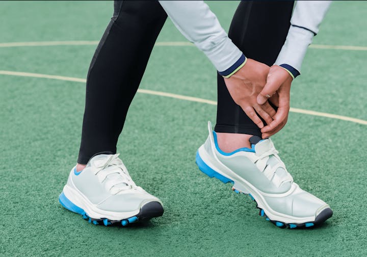 Person holding their ankle while standing on a green surface, wearing sports shoes and black leggings.