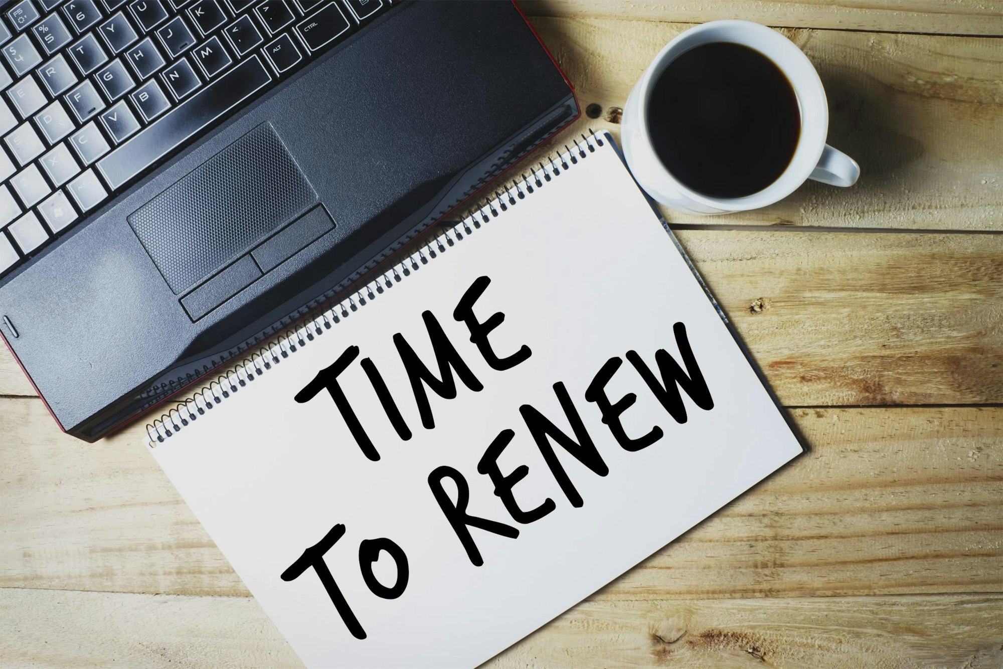 Laptop, notepad with "TIME To RENEW" written on it, and a cup of coffee on a wooden desk.