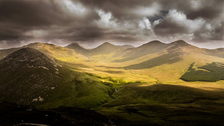 Mountain range with moody skies and patches of sunlight on the rolling green landscape.