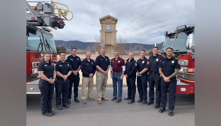 Group of firefighters and officials standing in front of a fire truck with a scenic mountain backdrop.