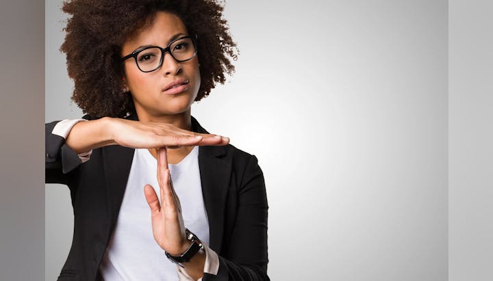 A person gesturing "timeout" or pausing, wearing glasses and a black blazer over a white top, with a neutral background.