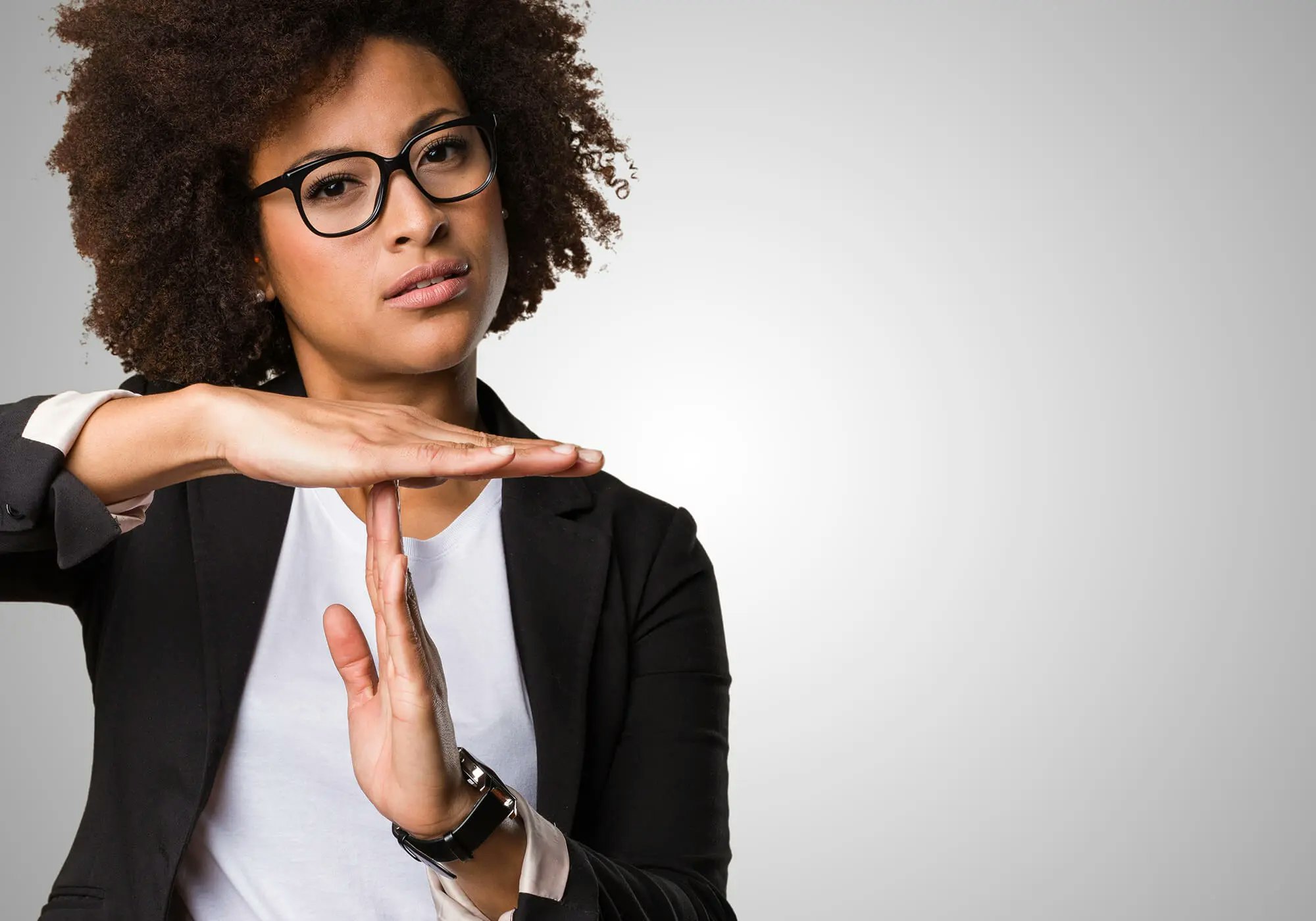 A person gesturing "timeout" or pausing, wearing glasses and a black blazer over a white top, with a neutral background.