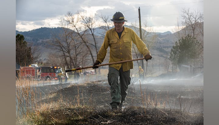 A firefighter with a tool stands before a smokey area with fire trucks in the background.