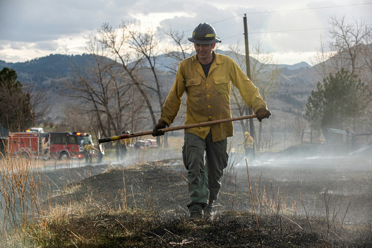 A firefighter with a tool stands before a smokey area with fire trucks in the background.