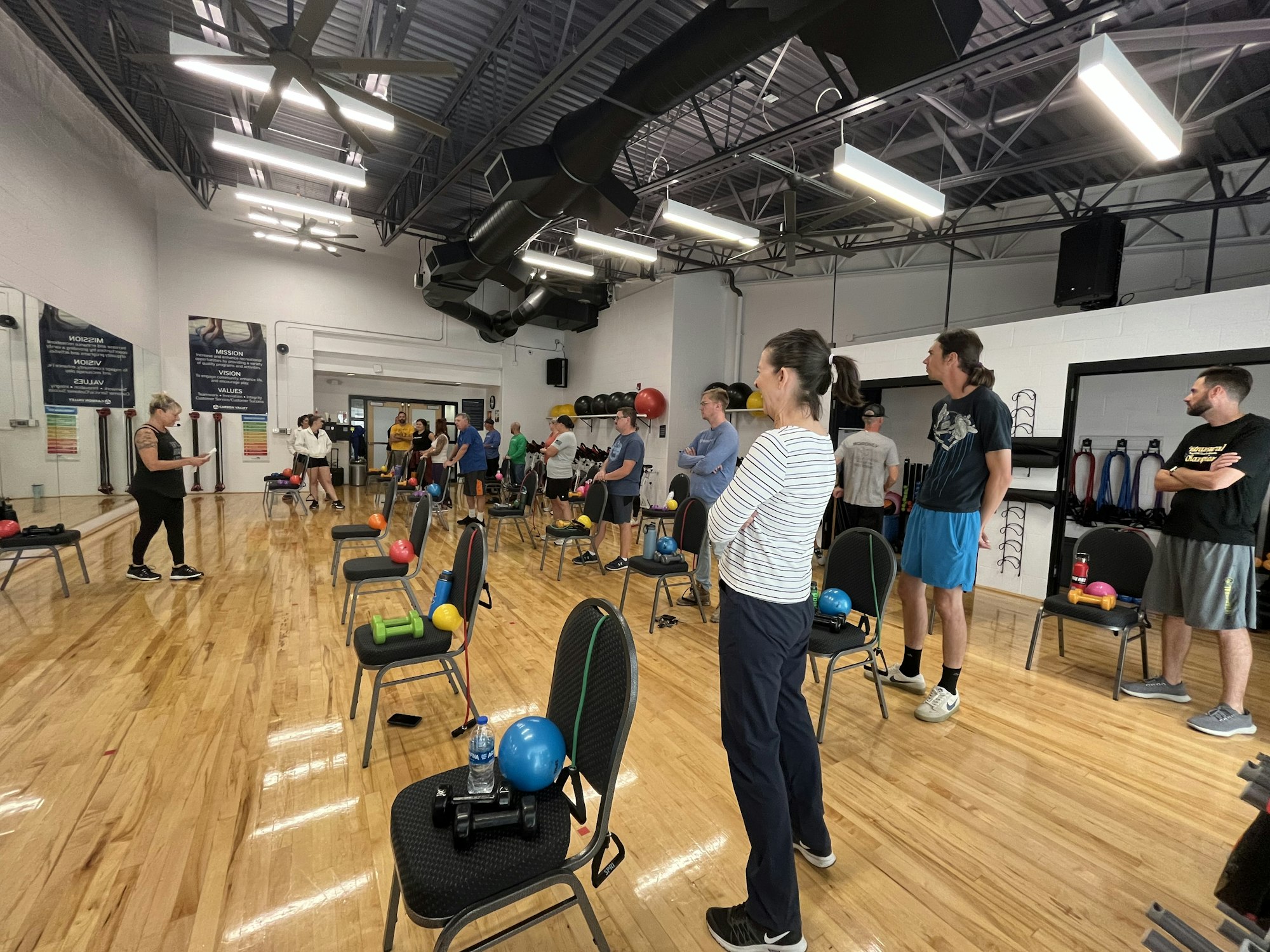 A group fitness class with people standing by chairs holding exercise equipment in a gym studio.