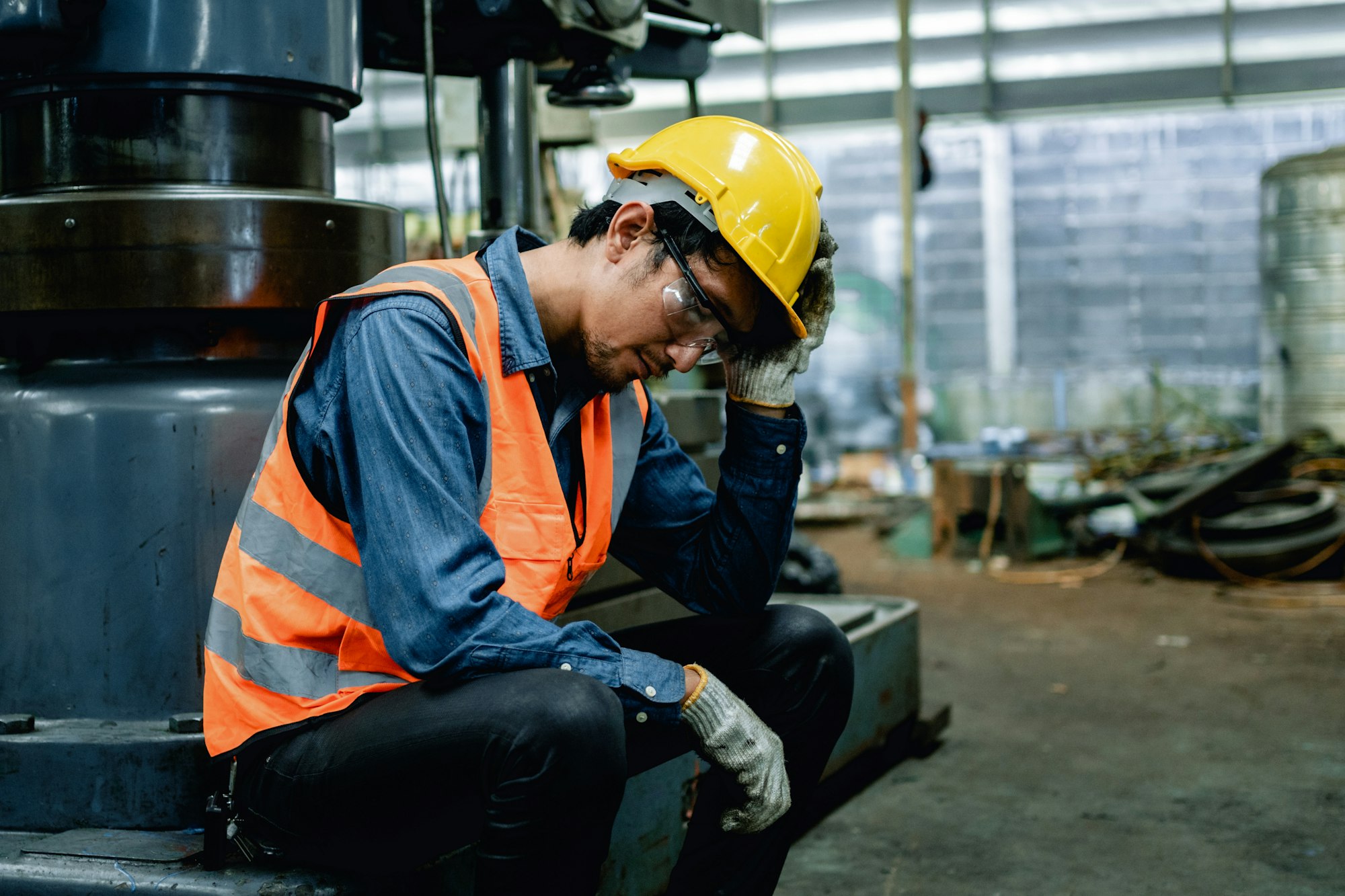 A worker in safety gear looks stressed while sitting on machinery in an industrial setting.
