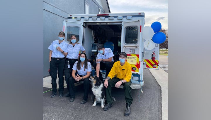 Five emergency responders and a dog in front of an ambulance with balloons.