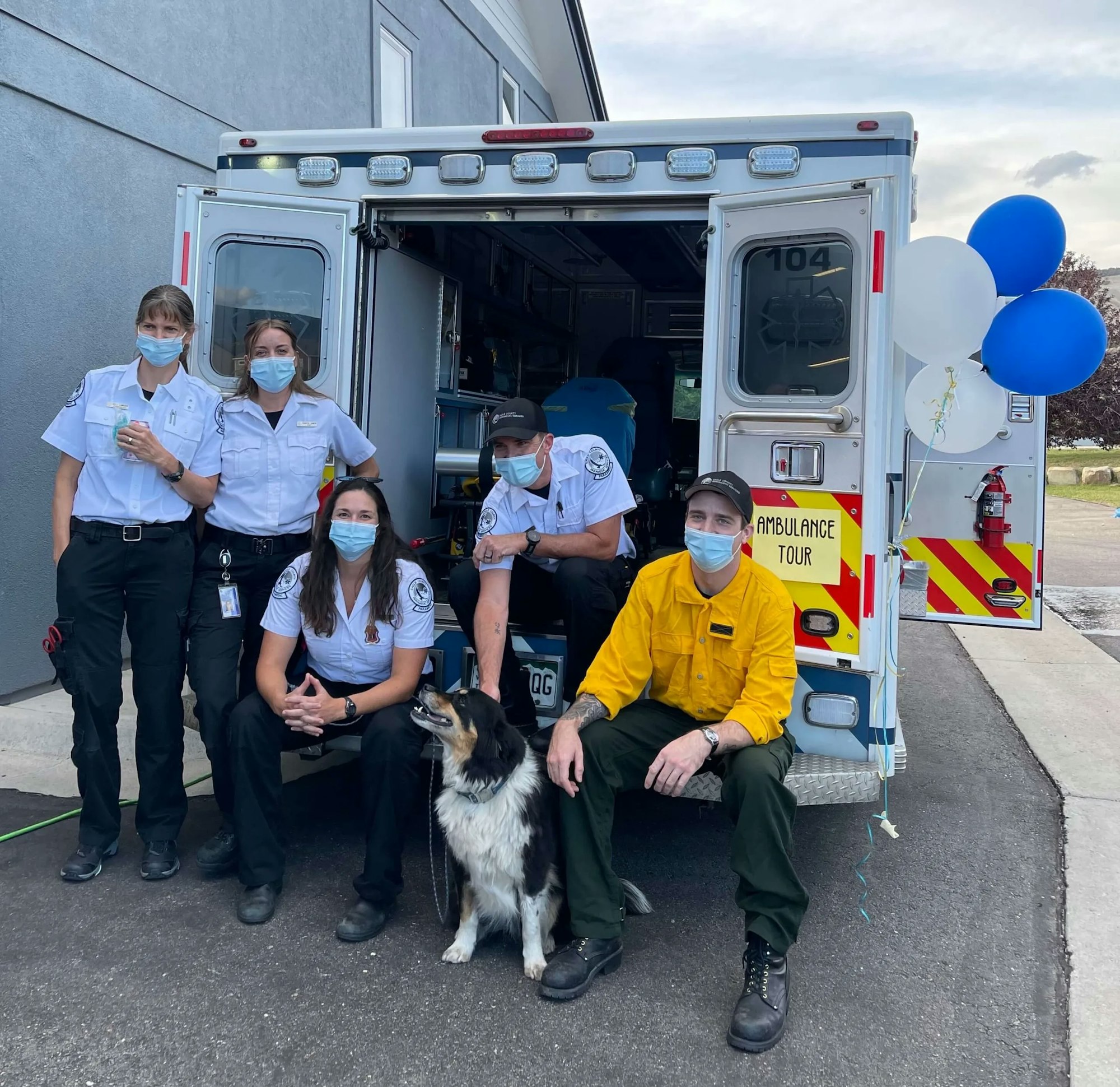 Five emergency responders and a dog in front of an ambulance with balloons.