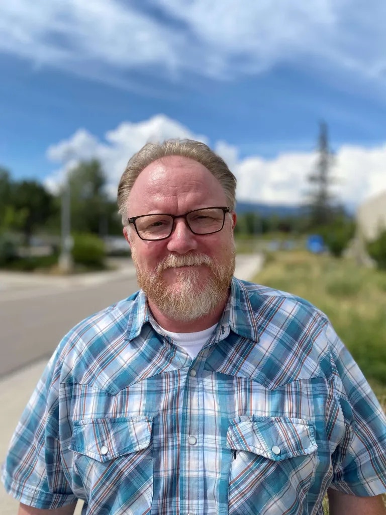 A smiling man with glasses, a beard, and a plaid shirt, outdoors with a blue sky and clouds in the background.