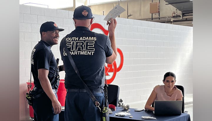 Two firefighters and a woman at a desk with a laptop.