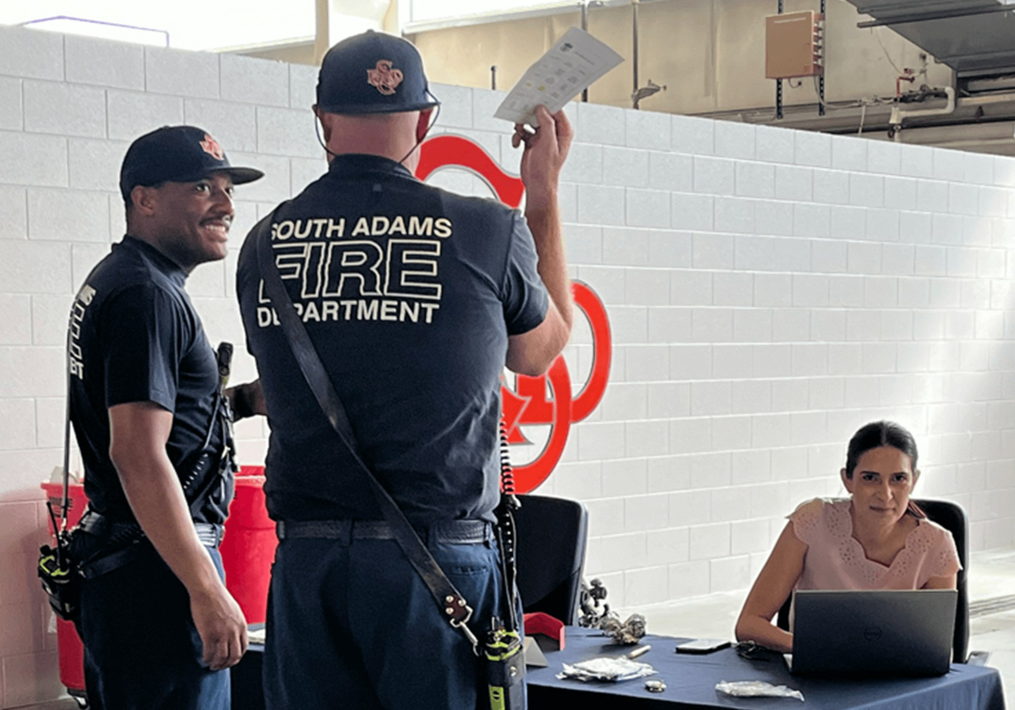 Two firefighters and a woman at a desk with a laptop.