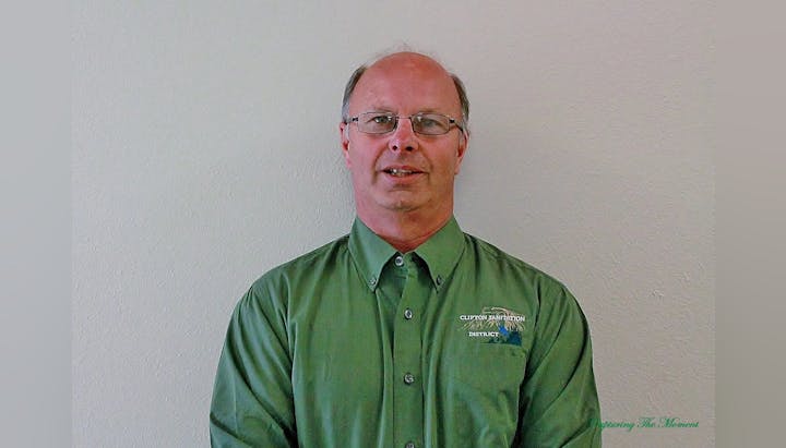 A man in a green shirt standing against a plain wall.