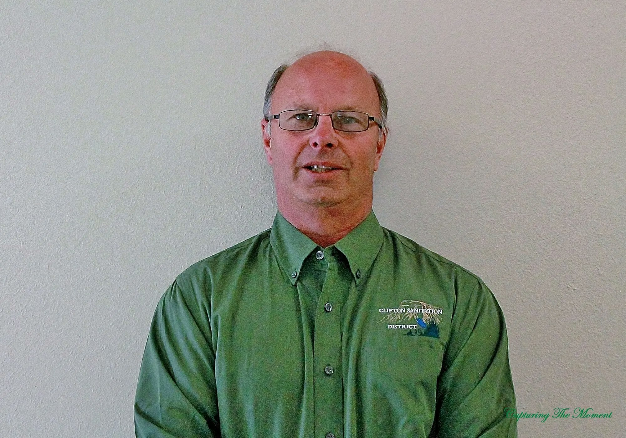A man in a green shirt standing against a plain wall.
