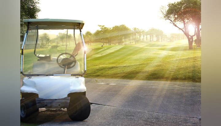 A golf cart in the foreground with a sunlit golf course and trees in the background.