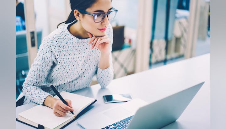 A woman wearing glasses is writing in a notebook while looking at a laptop in a bright, modern workspace.