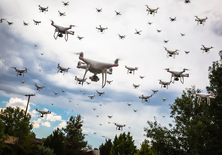 A swarm of drones flying under a cloudy sky, above some trees and a utility pole.