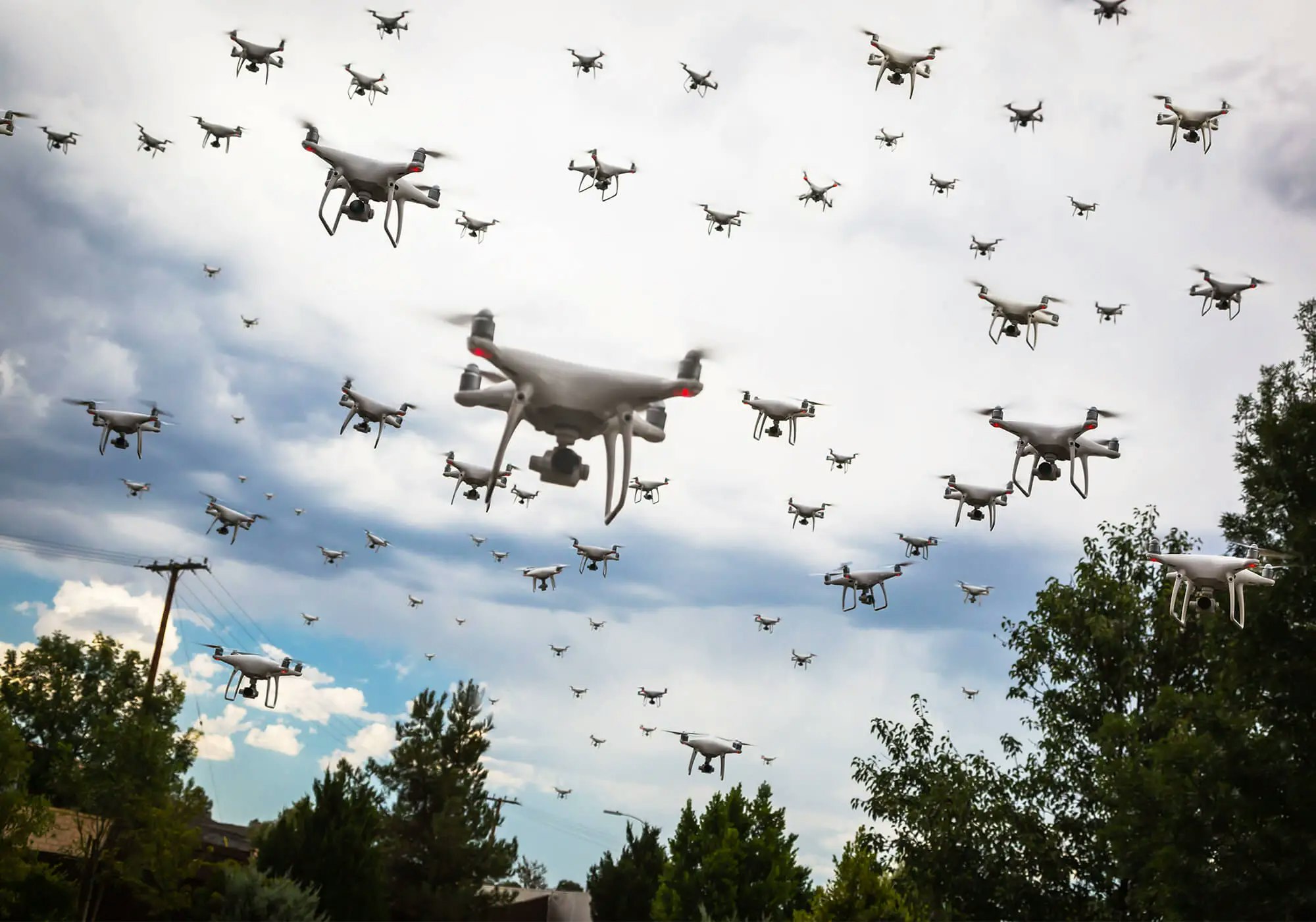 A swarm of drones flying under a cloudy sky, above some trees and a utility pole.