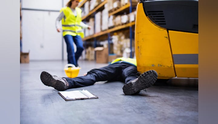 A worker lying on the floor of a warehouse with another person approaching; an industrial accident appears to have occurred.