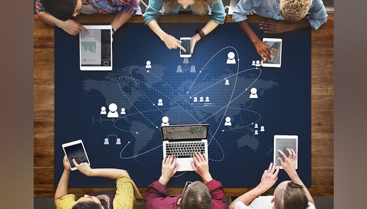 Group of people using smartphones, tablets, and laptop on a table with a global network graphic overlay.