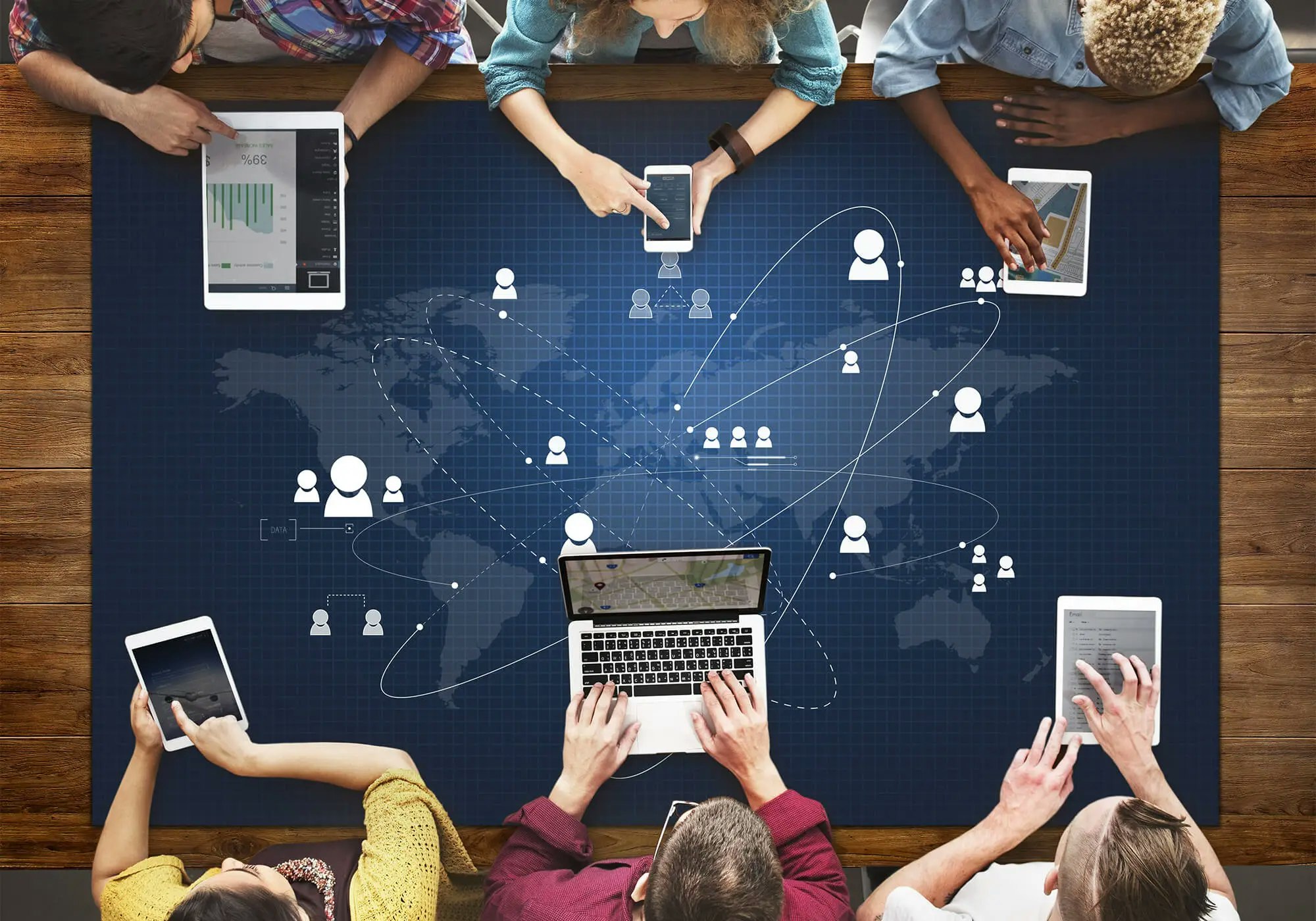 Group of people using smartphones, tablets, and laptop on a table with a global network graphic overlay.