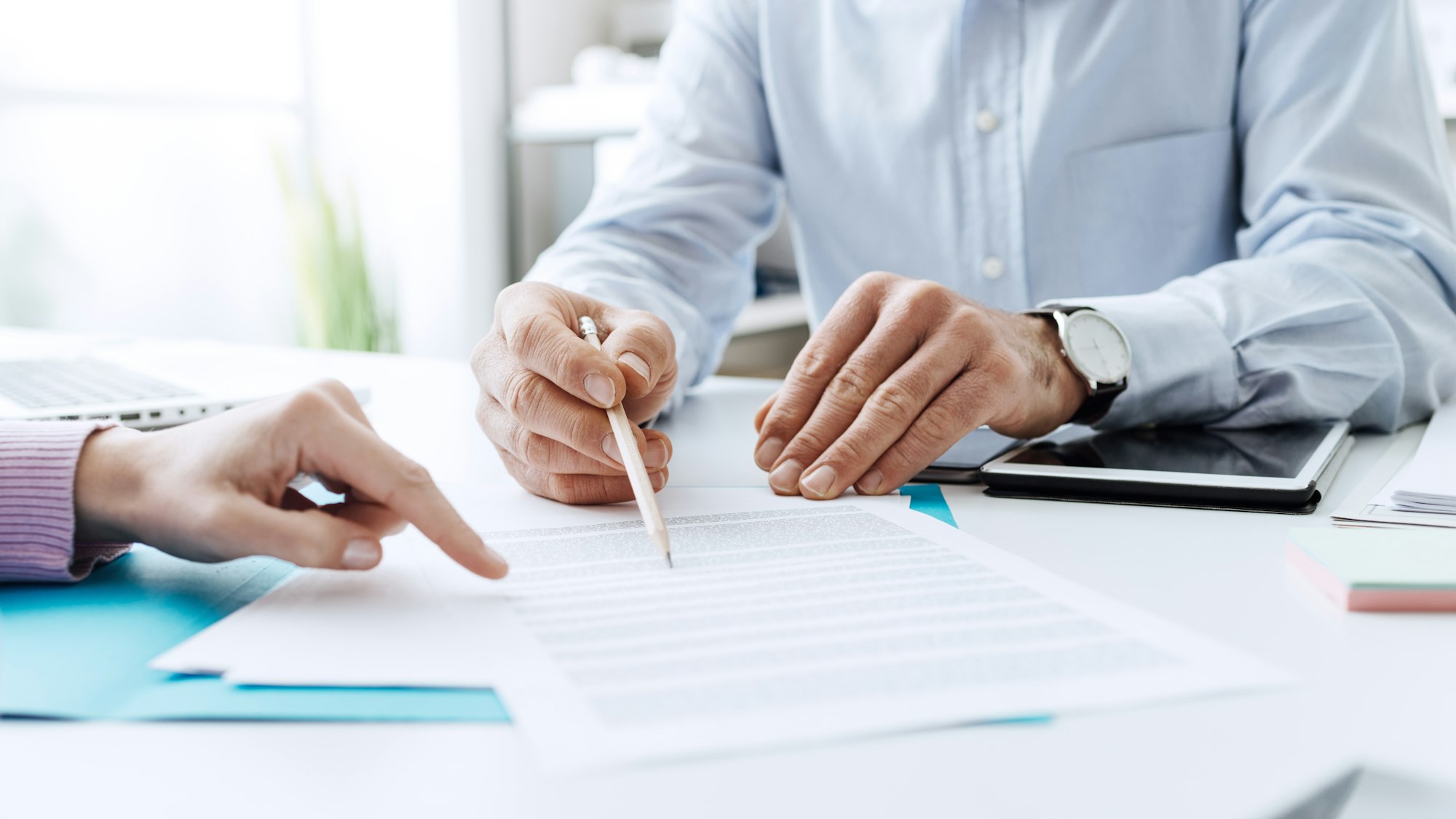 Two people discussing a document at a desk, one pointing at the paper while the other holds a pencil. A laptop and tablet are nearby.