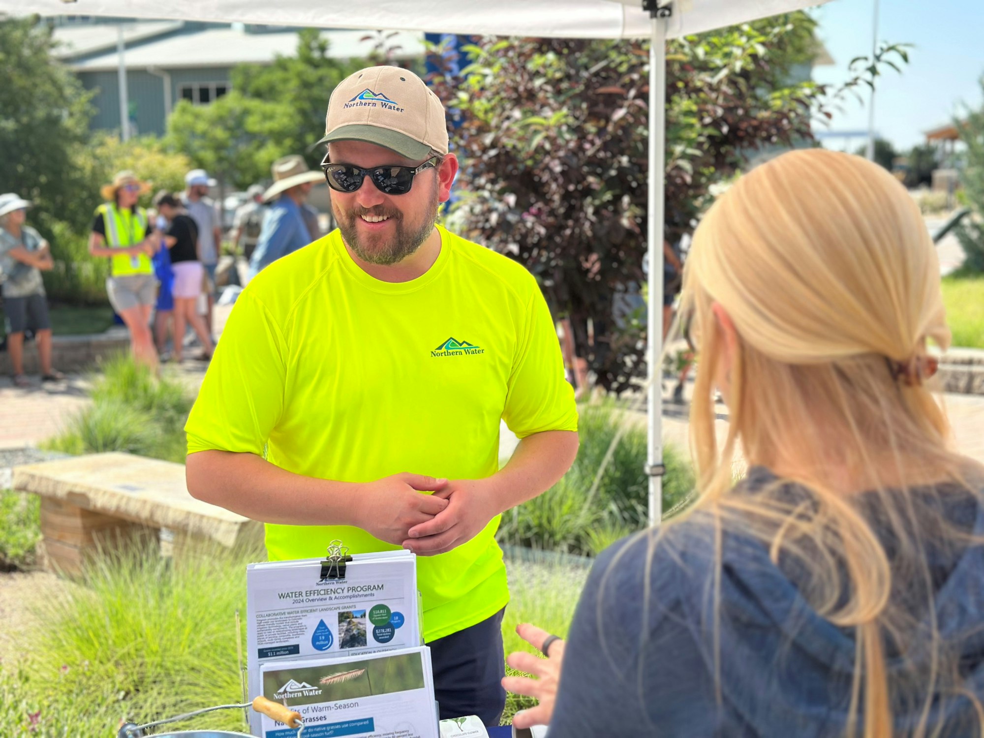 A man in a bright shirt speaks to a woman at a water efficiency program event, with others in the background.