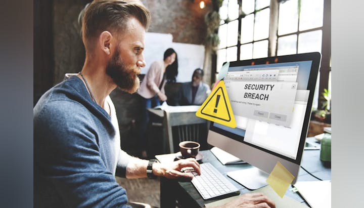 Man at desk with computer displaying a 'Security Breach' warning; colleagues in background.