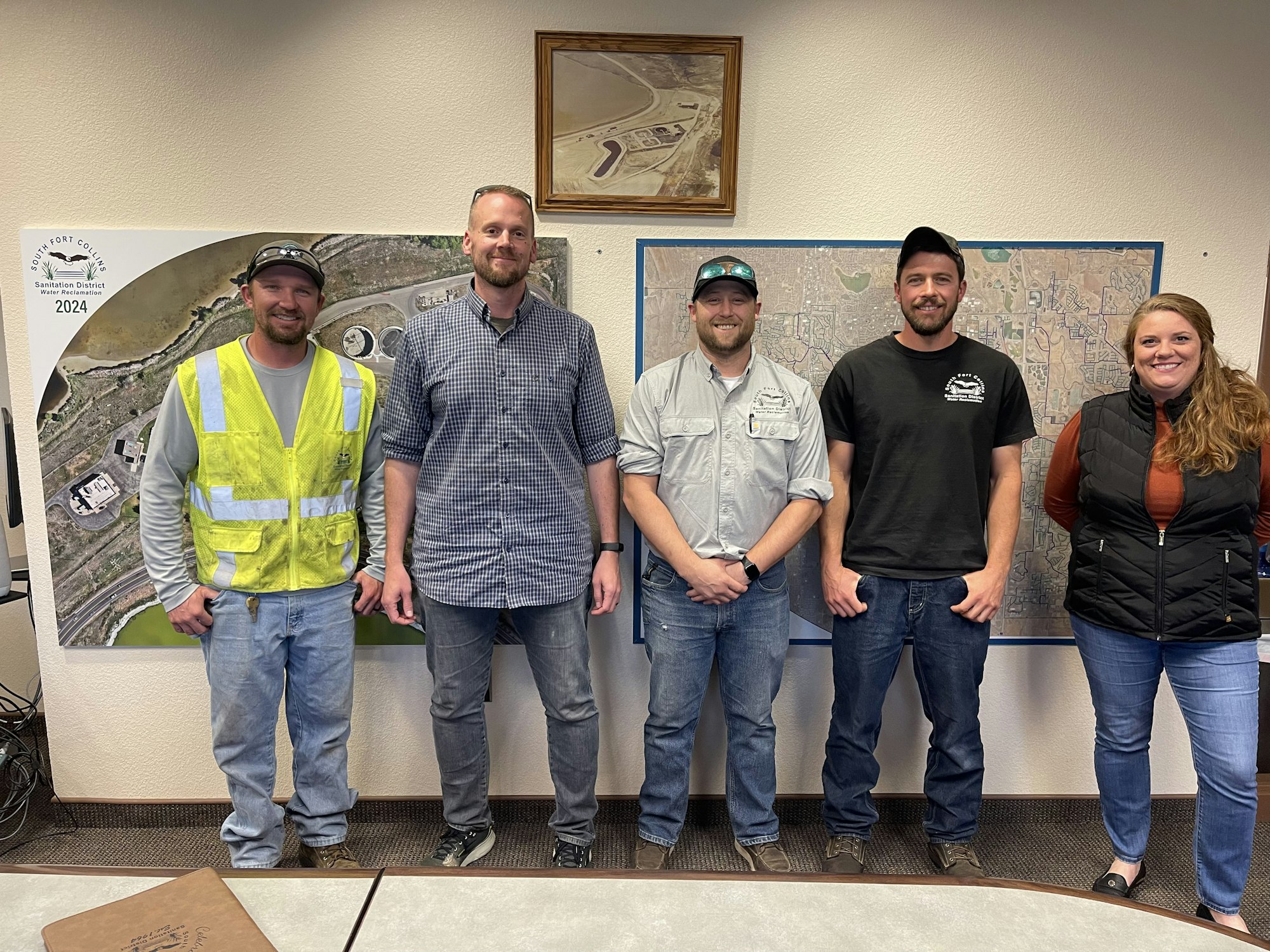 Five people standing in front of maps, some wearing casual clothing, one in a safety vest.