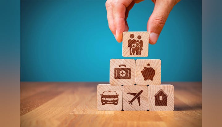 A hand placing a wooden block with a family icon atop a pyramid of blocks showing health, savings, a car, travel, and home.