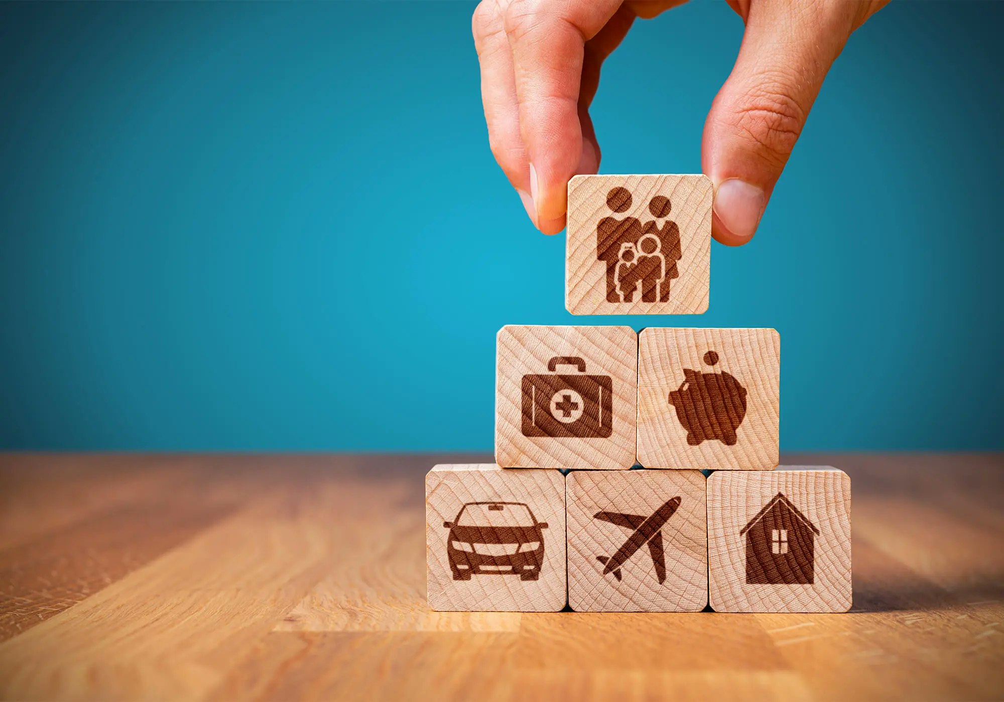 A hand placing a wooden block with a family icon atop a pyramid of blocks showing health, savings, a car, travel, and home.