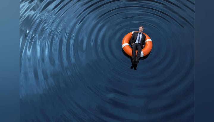 A person in a suit seated on an orange lifebuoy in the middle of rippling blue water.