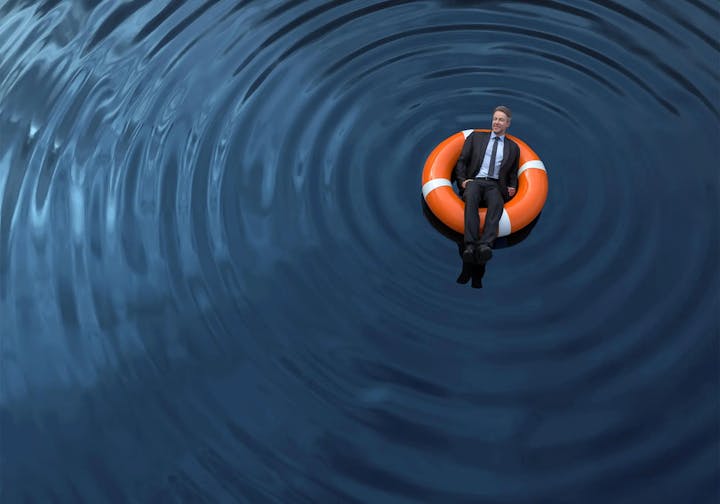 A person in a suit seated on an orange lifebuoy in the middle of rippling blue water.