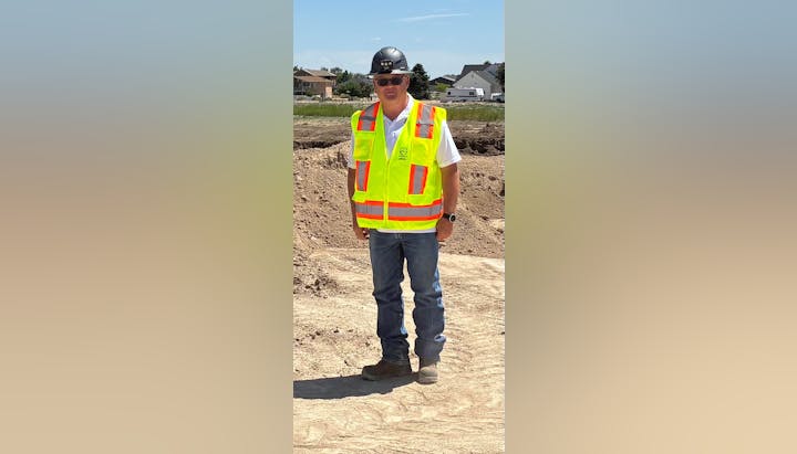 A person in safety gear and a hard hat stands on a construction site with sandy soil, wearing a bright reflective vest and sunglasses.