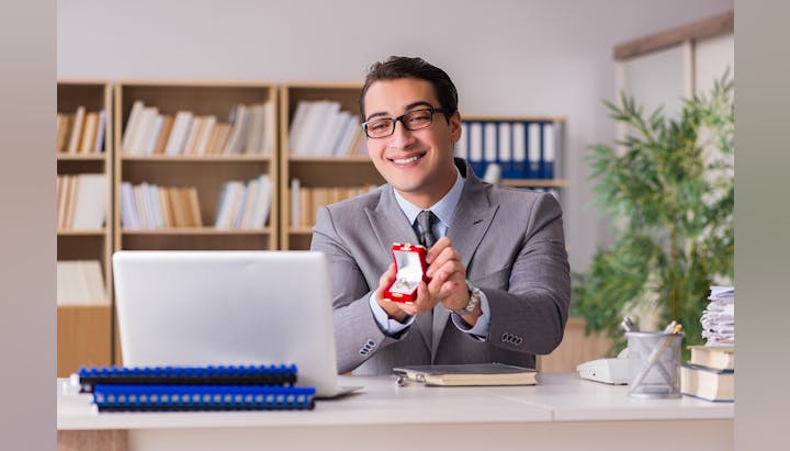 Man in suit holding a ring box with a smile at his desk.