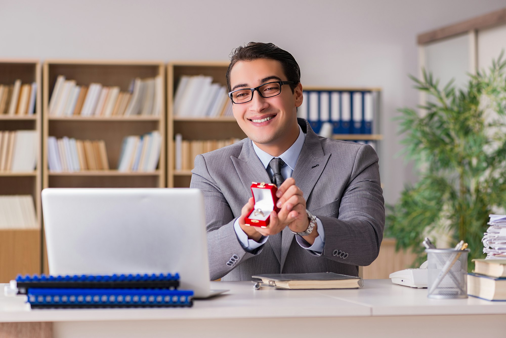Man in suit holding a ring box with a smile at his desk.