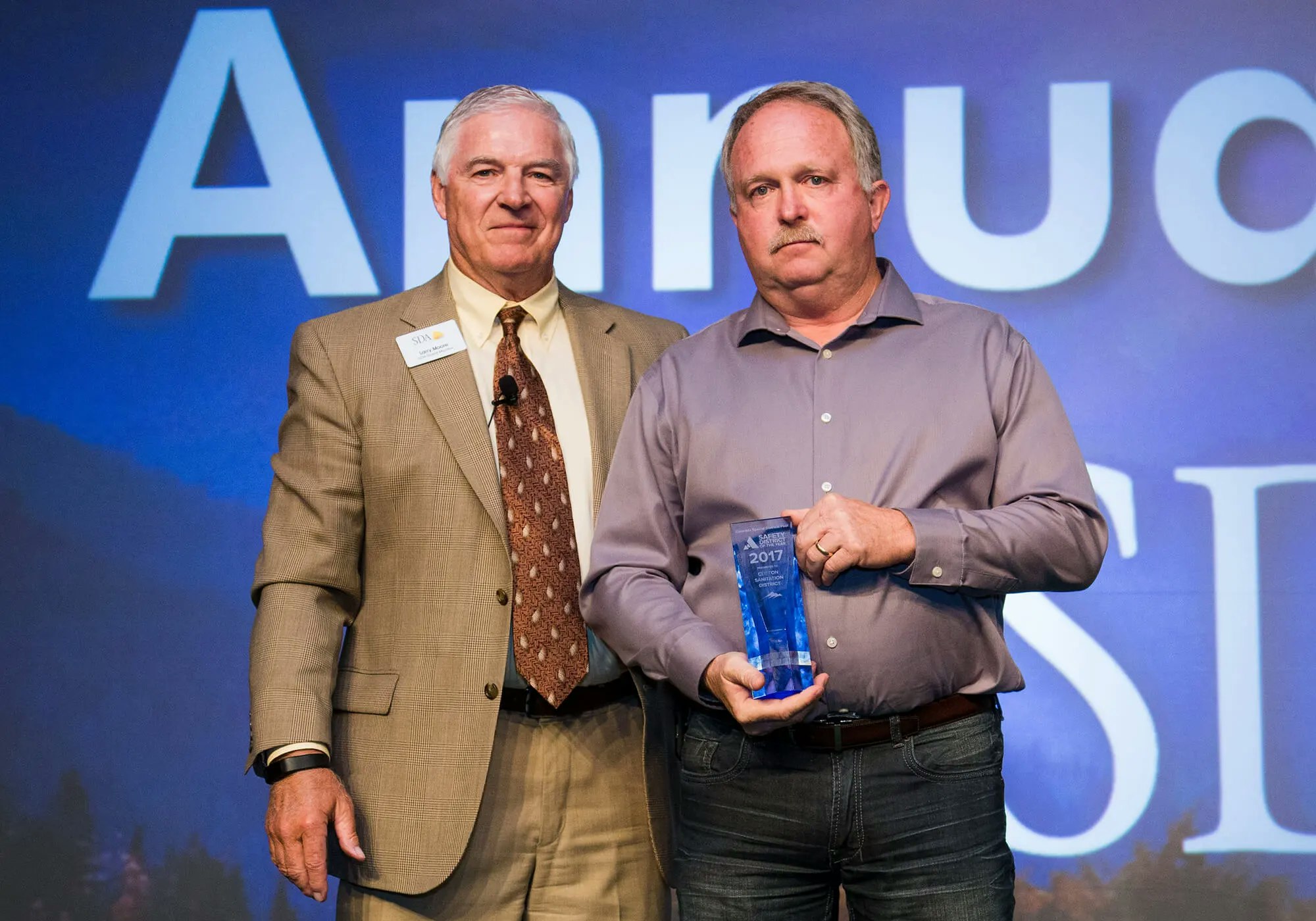 Two men on stage, one holding an award, with "AWARD" text in the background.