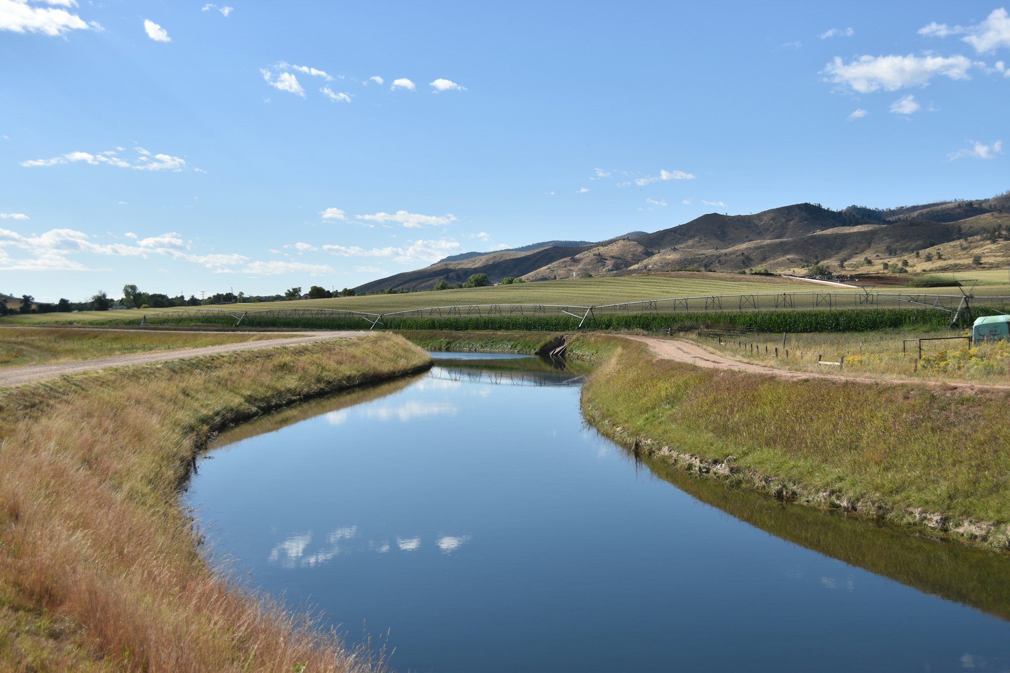 A winding waterway reflects blue skies, bordered by green fields and rolling hills under a sunny atmosphere.