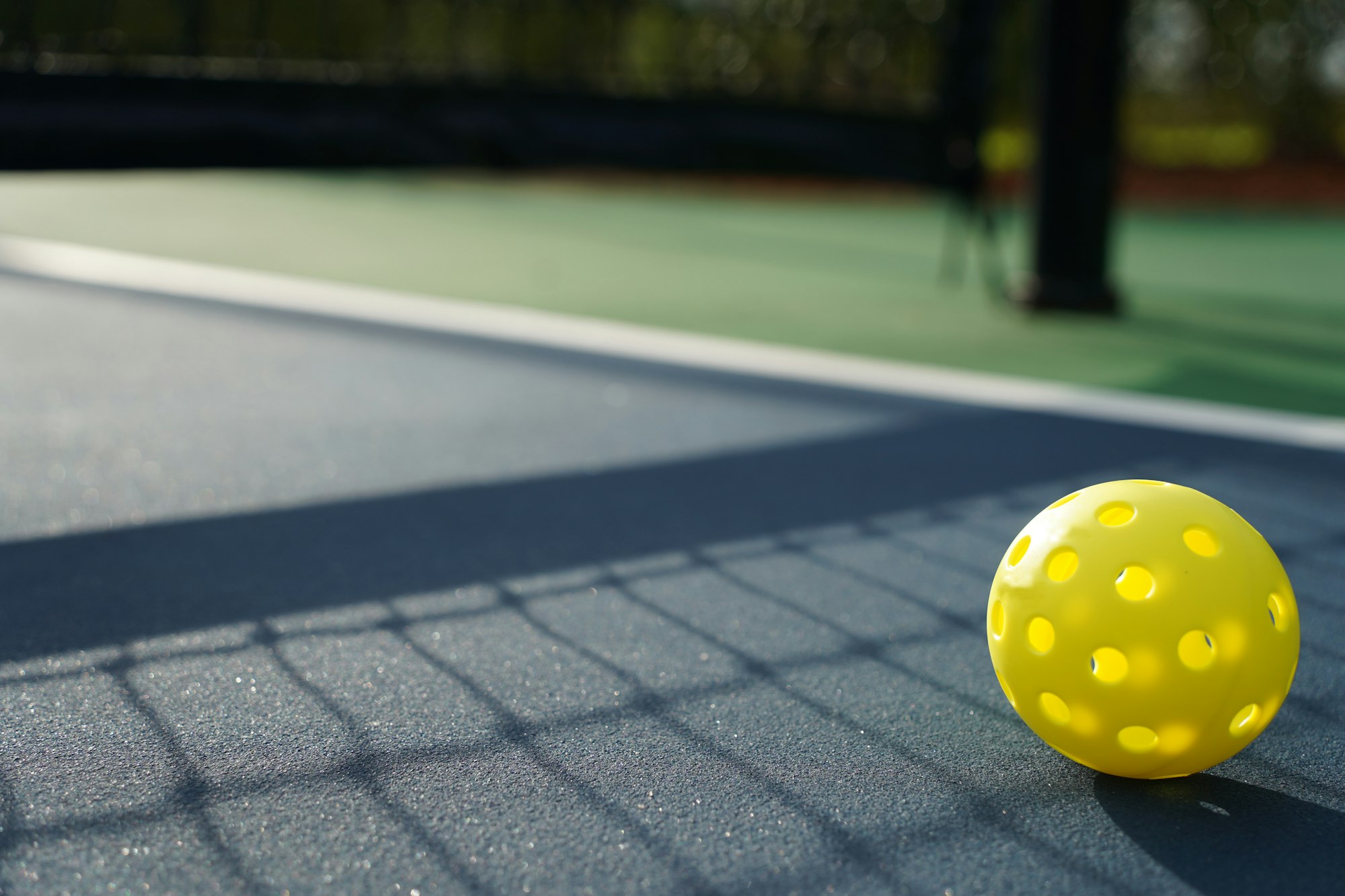 A close-up of a yellow perforated ball resting on a court surface, with shadows from a nearby net.