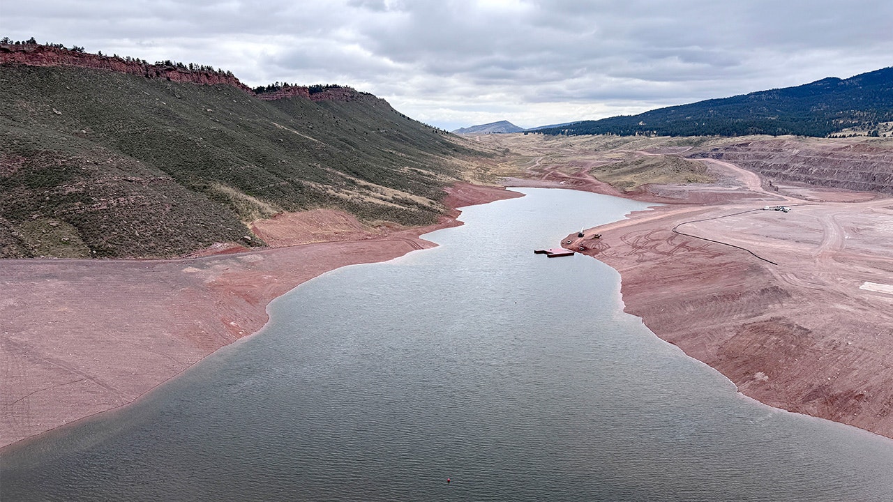 A river flows through a valley with reddish-brown terrain and green hills, under a cloudy sky. Heavy machinery is visible nearby.