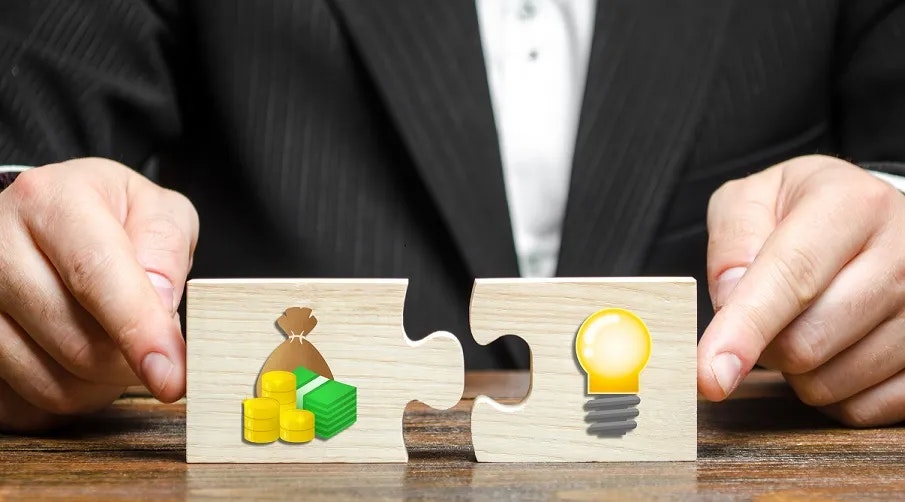 Two hands connecting puzzle pieces, one with money symbols, the other with a light bulb, suggesting a concept of funding or investing in ideas.