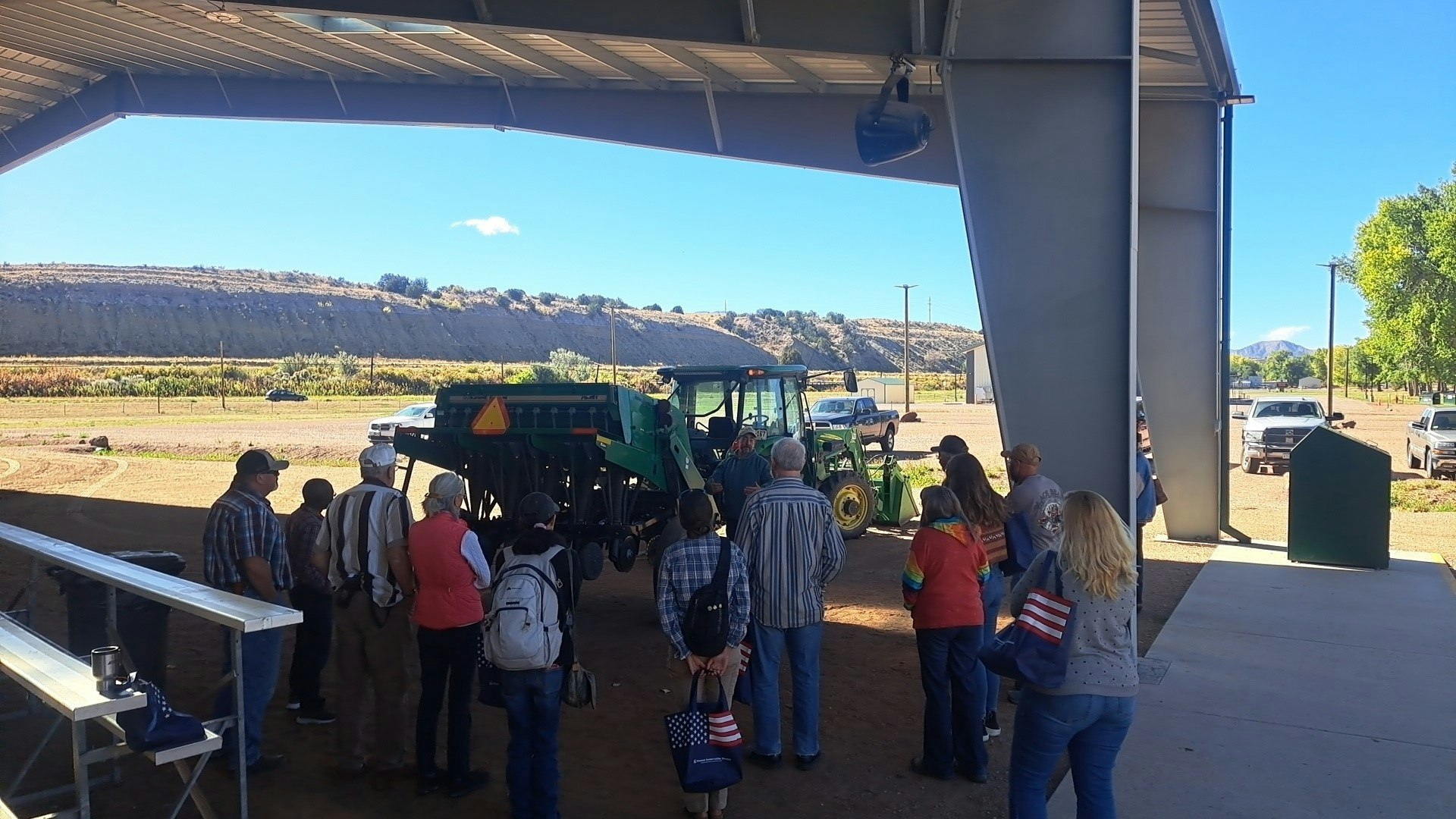A group of people gathers around a tractor under a shelter, with hills and vehicles in the background on a sunny day.