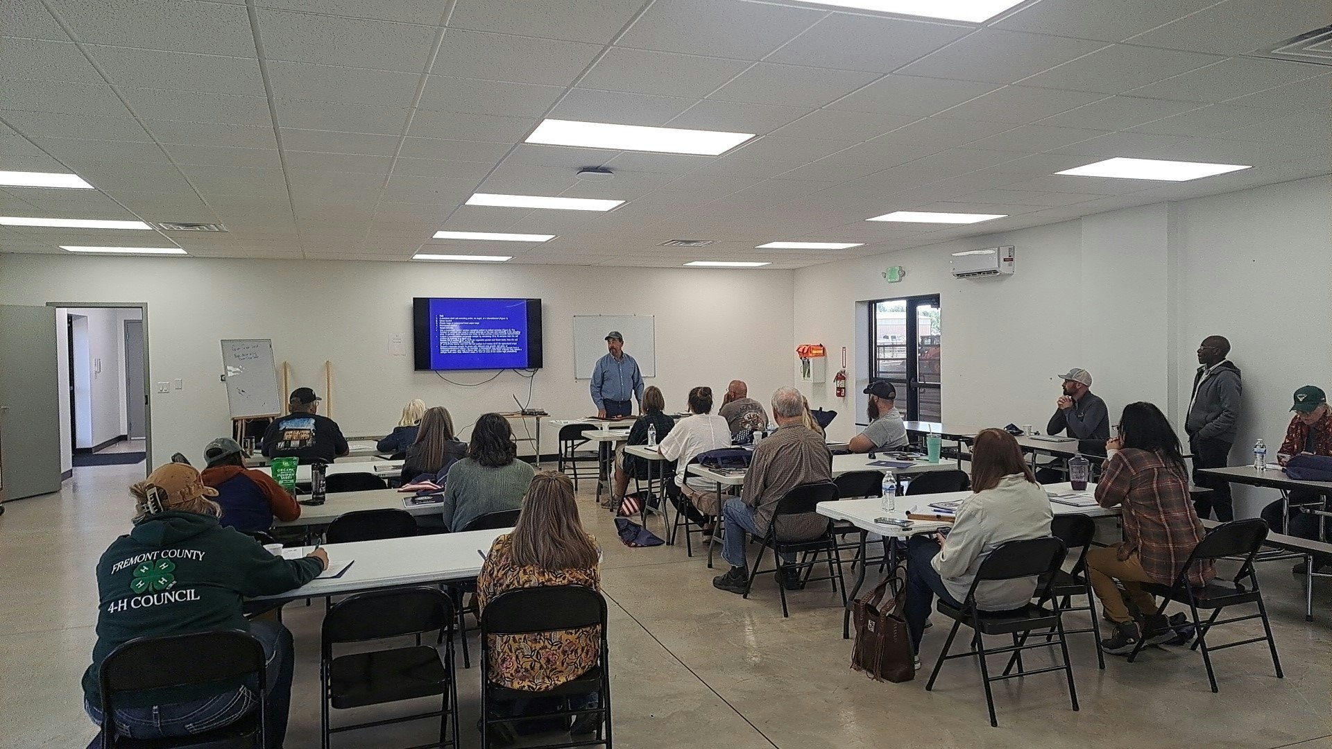 A group of people attending a meeting or presentation in a classroom setting, with a presenter and a screen displaying information.