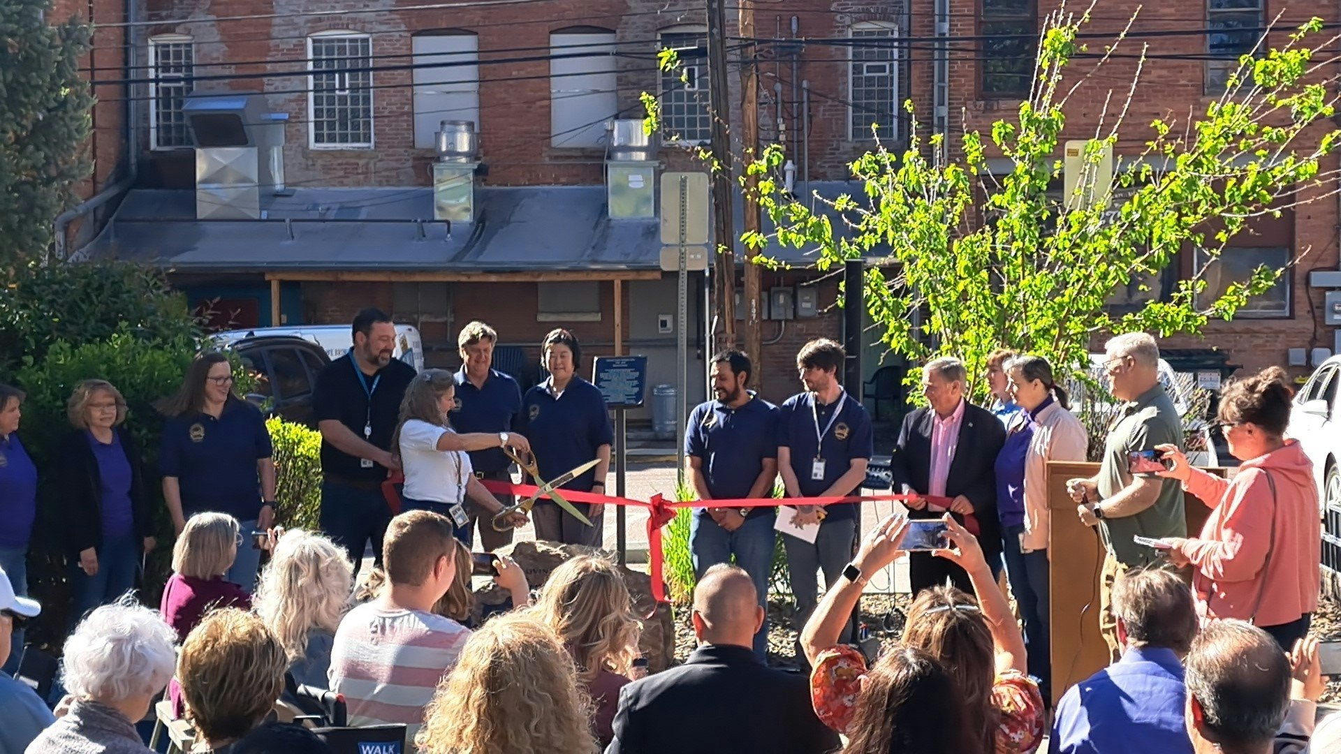 A group of people gathered for a ribbon-cutting ceremony outdoors, with a crowd watching.
